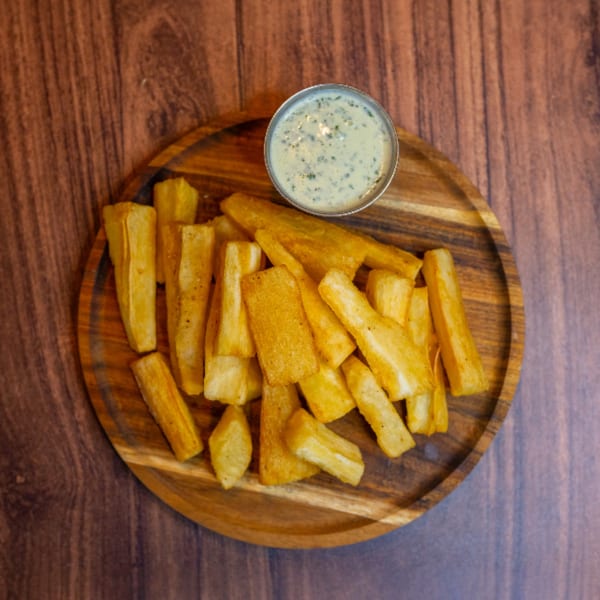 Golden fried yuca fries with a side of creamy green dipping sauce on a wooden plate.