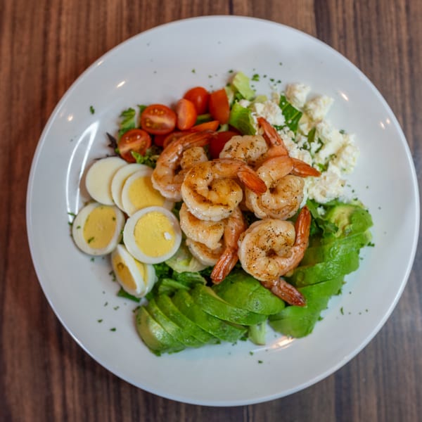 A vibrant shrimp salad with avocado, cherry tomatoes, hard-boiled eggs, and feta cheese on a white plate.